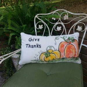 May include: A white pillow with the words "Give Thanks" and a hand-painted design of three pumpkins. The pumpkins are orange, yellow, and green. The pillow is sitting on a green chair.