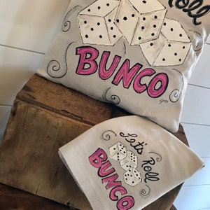 Peut inclure: Un coussin gris clair et une serviette carr&eacute;e assortie avec un motif de d&eacute;s et de texte. Le mot "Bunco" est en rose, et l'expression "Let's Roll" est en noir. Les d&eacute;s sont blancs avec des points noirs.