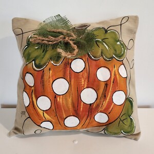 Puede incluir: Una almohada beige con una calabaza naranja pintada a mano con lunares blancos. La calabaza tiene hojas verdes y un lazo de arpillera marr&oacute;n con un lazo de malla verde en la parte superior.