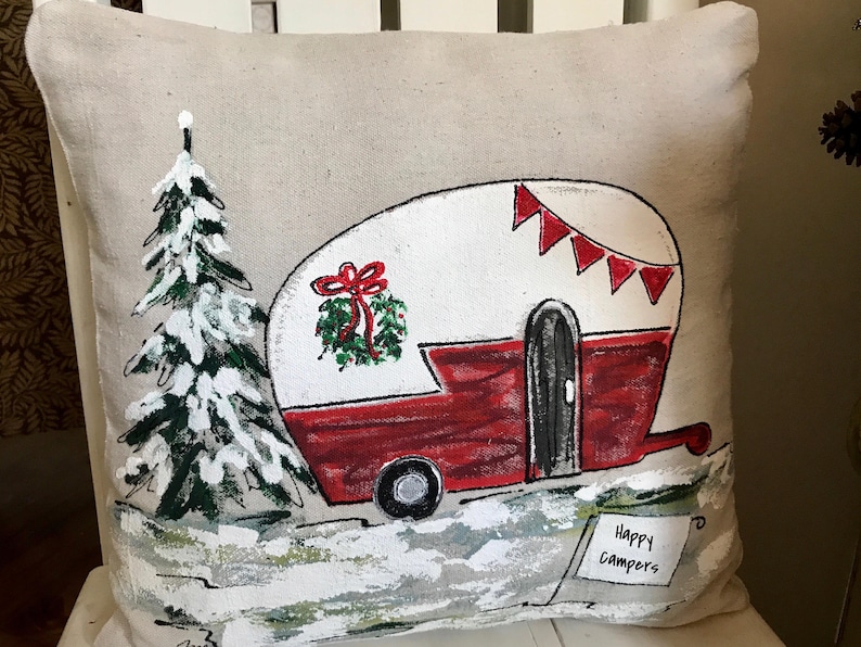Puede incluir: Una almohada beige con un dise&ntilde;o pintado a mano de una caravana roja con techo blanco y una corona verde en la parte delantera. La caravana tiene una puerta negra y una rueda negra. La caravana est&aacute; rodeada de un paisaje blanco nevado con un pino verde a la izquierda. La almohada tiene el texto "Happy Campers" escrito en un cartel blanco en la esquina inferior derecha.