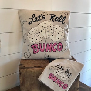 Peut inclure: Un coussin beige et un torchon assorti avec le texte "Let's Roll Bunco" en &eacute;criture noire et lettres roses. Trois d&eacute;s blancs avec des points noirs sont repr&eacute;sent&eacute;s. Le coussin est carr&eacute; et le torchon est rectangulaire.