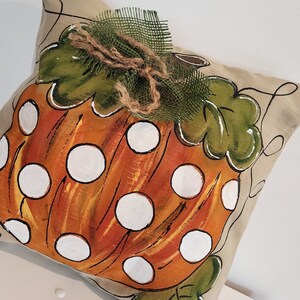Puede incluir: Una almohada beige con una calabaza naranja pintada a mano con lunares blancos. La calabaza tiene hojas verdes y un lazo de arpillera marr&oacute;n.
