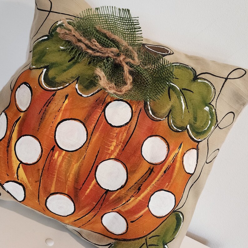 Puede incluir: Una almohada beige con una calabaza naranja pintada a mano con lunares blancos. La calabaza tiene hojas verdes y un lazo de arpillera marr&oacute;n.