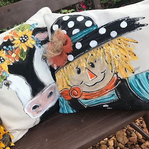 May include: Two decorative throw pillows with painted designs. One pillow features a black and white cow with a floral crown. The other pillow features a scarecrow with a black and white polka dot hat and a blue and white striped shirt.