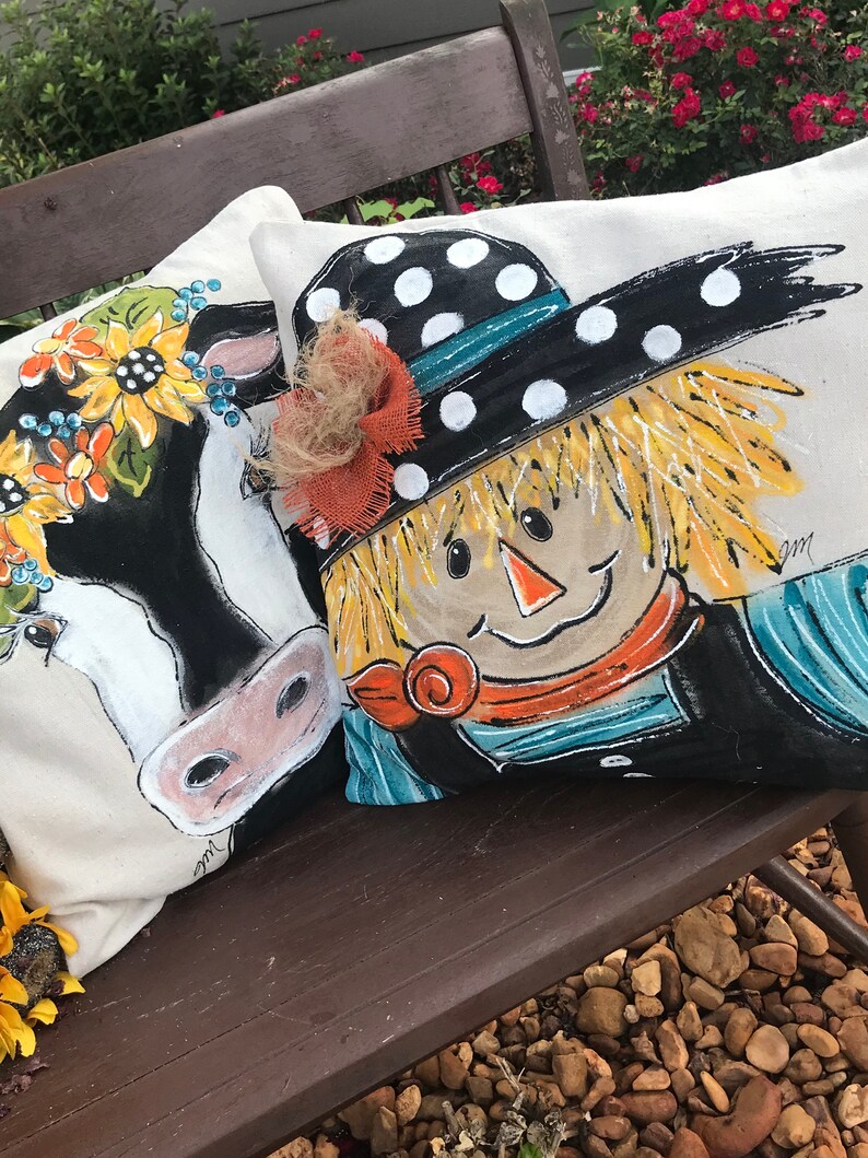 May include: Two decorative throw pillows with painted designs. One pillow features a black and white cow with a floral crown. The other pillow features a scarecrow with a black and white polka dot hat and a blue and white striped shirt.