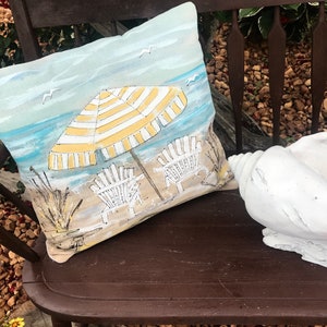 May include: A decorative cushion featuring a beach scene with a yellow and white striped umbrella, two white beach chairs, and seagulls flying overhead. The cushion is on a brown wooden bench.