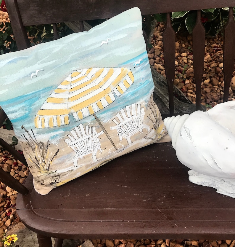 May include: A decorative cushion featuring a beach scene with a yellow and white striped umbrella, two white beach chairs, and seagulls flying overhead. The cushion is on a brown wooden bench.