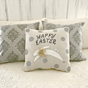 May include: A white pillow with gray polka dots and a white bunny with a flower on its head. The pillow has the text "Happy Easter" embroidered on it.