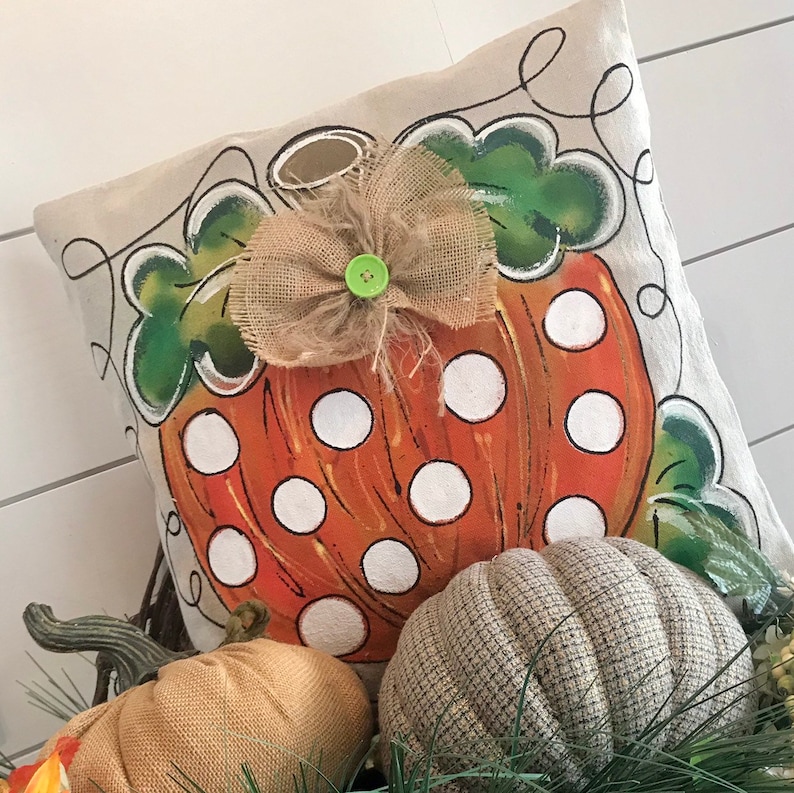 May include: A decorative pillow with a painted pumpkin design. The pumpkin is orange with white polka dots and has a burlap bow with a green button in the center. The pillow is surrounded by other pumpkins.