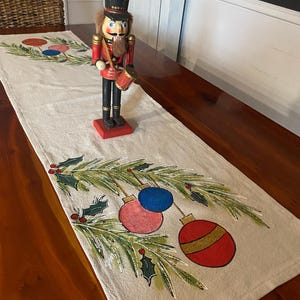 May include: A beige table runner with painted Christmas ornaments and greenery. A wooden nutcracker soldier with a drum stands on the runner. The runner features red, blue, and gold ornaments, and green holly.