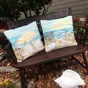 May include: Two decorative throw pillows with a beach theme. One pillow features a yellow and white striped umbrella with two beach chairs on a sandy beach. The other pillow features a seashell, sandcastles, and a sunset over the ocean.