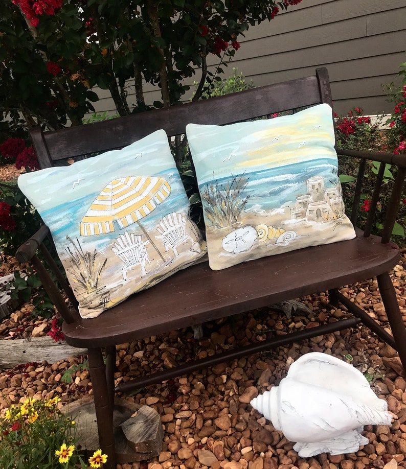 May include: Two decorative throw pillows with a beach theme. One pillow features a yellow and white striped umbrella with two beach chairs on a sandy beach. The other pillow features a seashell, sandcastles, and a sunset over the ocean.