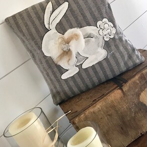 May include: A gray and white striped pillow with a white painted bunny rabbit with a white fluffy tail and a sparkly rhinestone embellishment. The pillow is sitting on a wooden box.