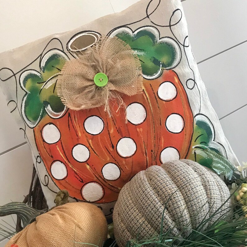 May include: A decorative pillow with a painted pumpkin design. The pumpkin is orange with white polka dots and has a green stem and leaves. A burlap bow with a green button is on top of the pumpkin. The pillow is on a white surface with other pumpkins.