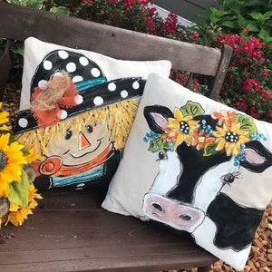 May include: Two decorative throw pillows with a farmhouse theme. One pillow features a painted scarecrow with a black and white polka dot hat and a red and orange scarf. The other pillow features a painted cow with a sunflower crown.