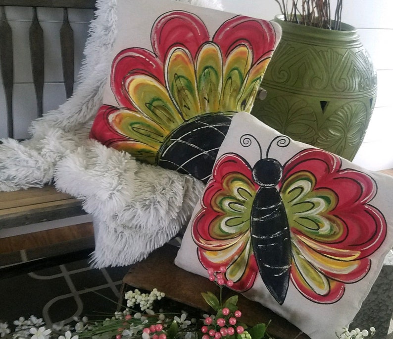 K&ouml;nnte beinhalten: Zwei dekorative Kissen mit Blumen- und Schmetterlingsmotiven. Das Blumenkissen hat rote, gr&uuml;ne und gelbe Akzente. Das Schmetterlingskissen hat rote, gr&uuml;ne, gelbe und schwarze Akzente.