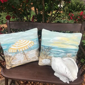 May include: Two decorative throw pillows with beach scenes on a wooden bench. One pillow features a yellow and white striped umbrella with two beach chairs on a sandy beach. The other pillow shows a seascape with a sunset and seagulls. A white seashell sits on the bench.
