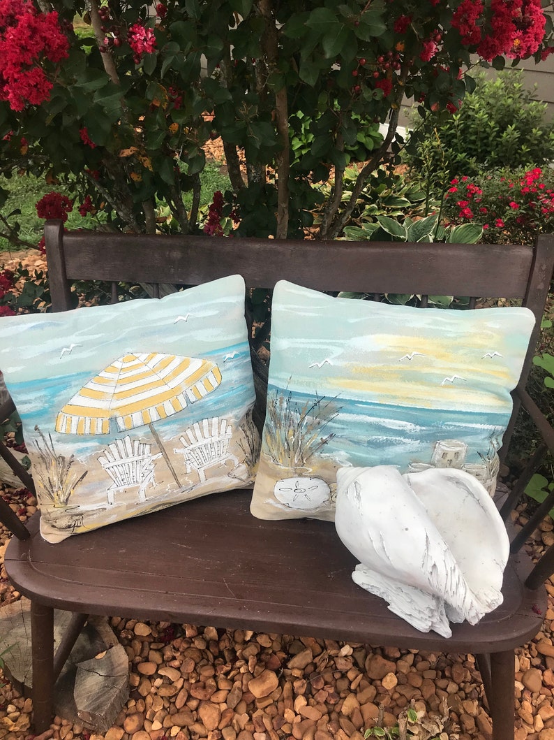 May include: Two decorative throw pillows with beach scenes on a wooden bench. One pillow features a yellow and white striped umbrella with two beach chairs on a sandy beach. The other pillow shows a seascape with a sunset and seagulls. A white seashell sits on the bench.