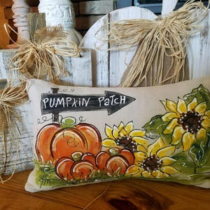 May include: A beige pillow with a painted design of pumpkins and sunflowers. The pillow has the words "Pumpkin Patch" written on a black sign.