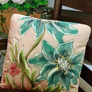 K&ouml;nnte beinhalten: Ein dekoratives Kissen mit einem Blumendesign mit gro&szlig;en, blaugr&uuml;nen Bl&uuml;ten und gr&uuml;nen Bl&auml;ttern auf einem hellrosa Hintergrund.