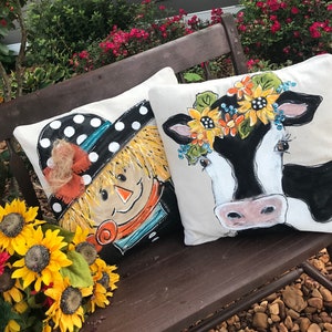 May include: Two decorative throw pillows with painted designs. One pillow features a scarecrow with a black and white polka dot hat and a straw brim. The other pillow features a black and white cow with a flower crown.