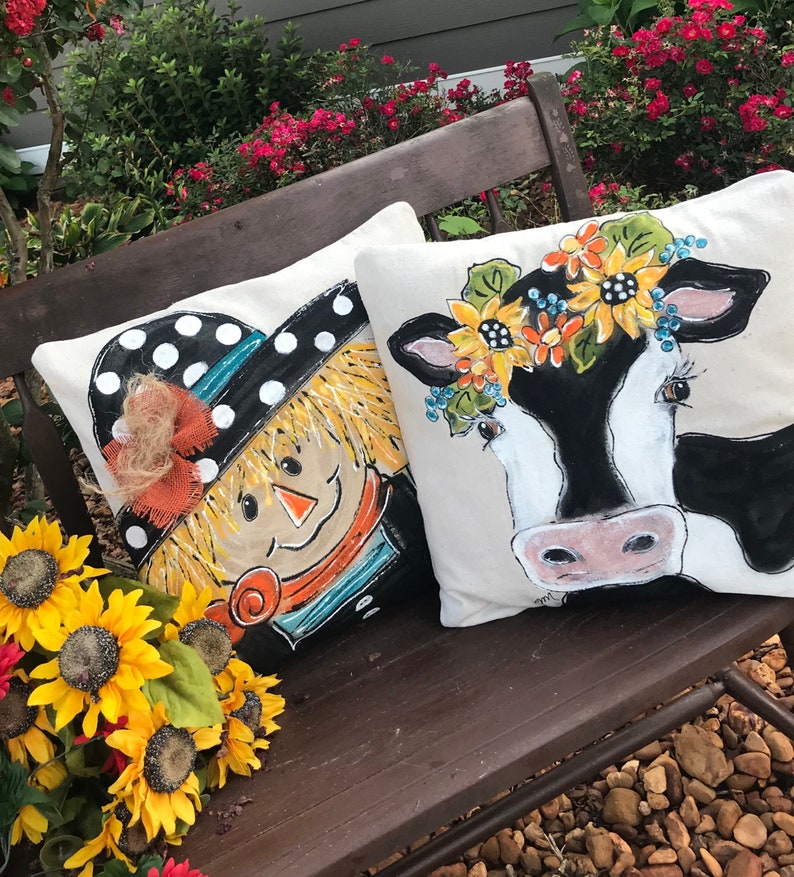 May include: Two decorative throw pillows with painted designs. One pillow features a scarecrow with a black and white polka dot hat and a straw brim. The other pillow features a black and white cow with a flower crown.