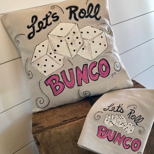 Peut inclure: Un ensemble coussin et torchon beiges avec le texte "Let's Roll Bunco" en noir et rose. Le motif pr&eacute;sente trois d&eacute;s blancs avec des points noirs. Le coussin est de forme carr&eacute;e et le torchon est rectangulaire.