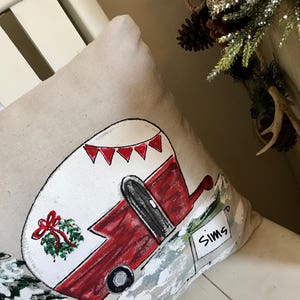 Puede incluir: Una almohada beige con un dise&ntilde;o pintado a mano de una caravana roja y blanca con una corona en el lateral. La caravana tiene un cartel que dice "Sims".