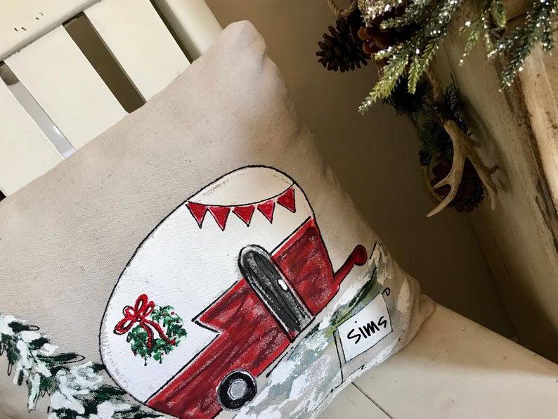Puede incluir: Una almohada beige con un dise&ntilde;o pintado a mano de una caravana roja y blanca con una corona en el lateral. La caravana tiene un cartel que dice "Sims".