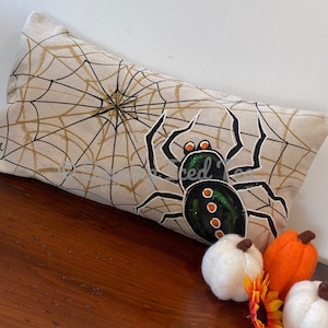 May include: A decorative pillow with a black spider on a white background with a gold glitter spider web. The spider has green and orange accents.