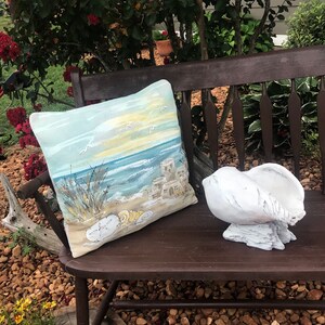 May include: A wooden bench with a blue and yellow cushion featuring a beach scene with seashells. A white seashell sits on the bench.
