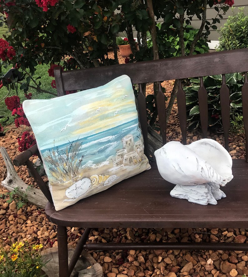May include: A wooden bench with a blue and yellow cushion featuring a beach scene with seashells. A white seashell sits on the bench.