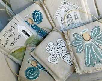 May include: Five handmade fabric ornaments with a rustic, farmhouse style. The ornaments feature a church, a cross, and angels with blue wings. The ornaments are made of natural linen fabric and have a frayed edge.