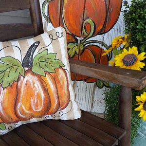 Pu&ograve; includere: Un cuscino decorativo con un designo di zucca dipinto su uno sfondo di tessuto a righe. Il cuscino &egrave; seduto su una sedia di legno con un cartello di zucca dipinto dietro.