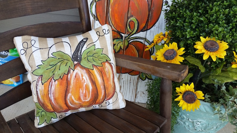 Pu&ograve; includere: Un cuscino decorativo con un designo di zucca dipinto su uno sfondo di tessuto a righe. Il cuscino &egrave; seduto su una sedia di legno con un cartello di zucca dipinto dietro.