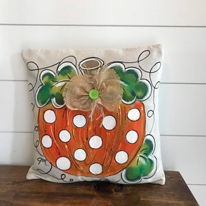 May include: A beige pillow with a hand-painted orange and white polka dot pumpkin design. The pumpkin has a green stem and a burlap bow with a green button in the center. The pillow has a white background and black outlines.