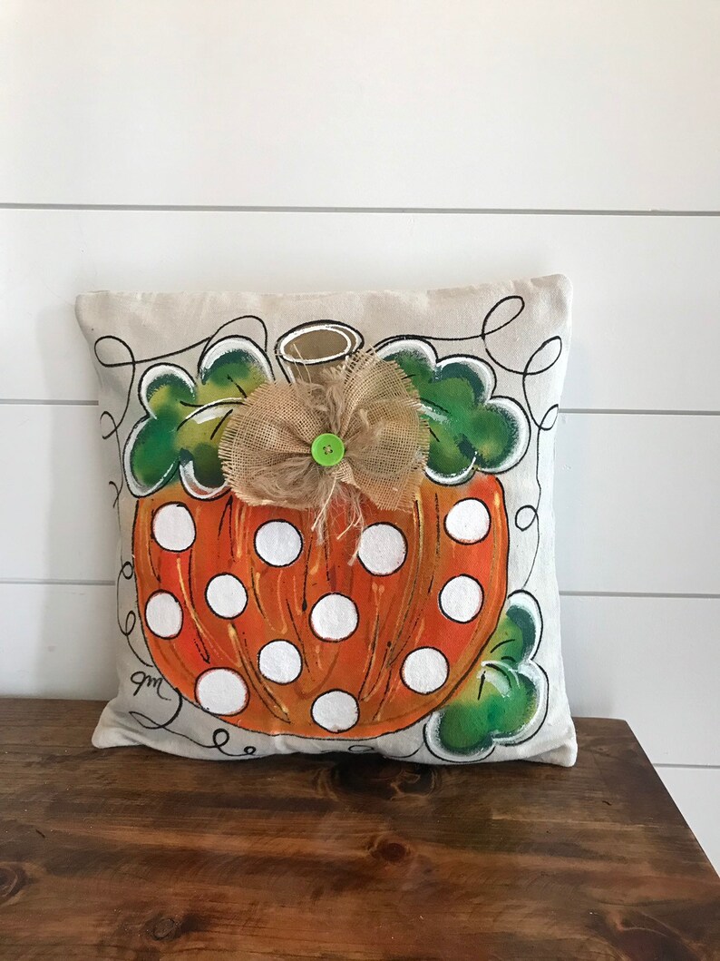 May include: A beige pillow with a hand-painted orange and white polka dot pumpkin design. The pumpkin has a green stem and a burlap bow with a green button in the center. The pillow has a white background and black outlines.