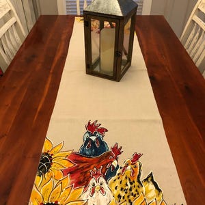 May include: A beige fabric table runner featuring a hand-painted design of three chickens and several bright yellow sunflowers. The chickens are depicted in shades of blue, red, white, and brown. A metal lantern sits on the runner.