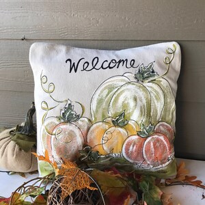 May include: A decorative pillow with a painted pumpkin design. The pillow features a variety of pumpkins in shades of orange, yellow, and green, with the word "Welcome" in black script. Autumn leaves and a woven ball complete the fall theme.