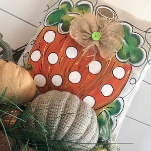 May include: A decorative pillow with a painted orange pumpkin with white polka dots and green leaves. The pumpkin has a burlap bow with a green button in the center.