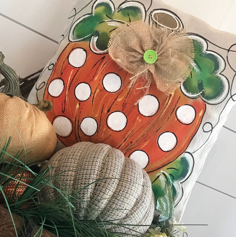 May include: A decorative pillow with a painted orange pumpkin with white polka dots and green leaves. The pumpkin has a burlap bow with a green button in the center.