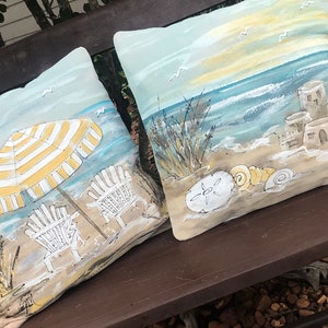 May include: Two decorative throw pillows with a beach theme. One pillow features a yellow and white striped umbrella, two beach chairs, and seagulls. The other pillow features a seashell, sandcastle, and ocean waves.