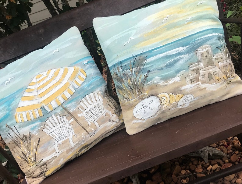 May include: Two decorative throw pillows with a beach theme. One pillow features a yellow and white striped umbrella, two beach chairs, and seagulls. The other pillow features a seashell, sandcastle, and ocean waves.