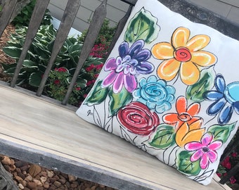 Peut inclure: Un oreiller blanc avec un motif floral coloré. Les fleurs sont peintes dans des couleurs vives, notamment le jaune, l'orange, le rouge, le rose, le bleu et le violet. L'oreiller est posé sur un banc en bois.