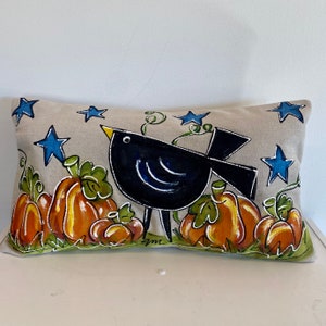 May include: A decorative pillow with a beige linen fabric features a hand-painted design of a black bird with blue accents standing amongst orange pumpkins and green leaves. The background is scattered with blue stars.