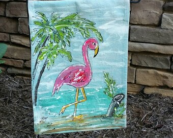 May include: A pink flamingo standing in blue water with a palm tree in the background. The flamingo is painted on a light blue fabric with a white border. The fabric is attached to a black metal pole.