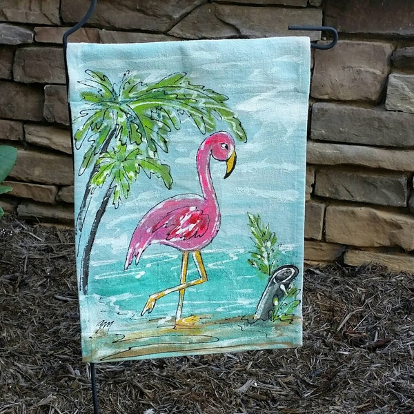 May include: A pink flamingo standing in blue water with a palm tree in the background. The flamingo is painted on a light blue fabric with a white border. The fabric is attached to a black metal pole.