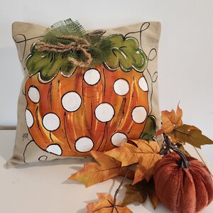 Puede incluir: Una almohada beige con un dise&ntilde;o de calabaza pintado a mano. La calabaza es naranja con lunares blancos y hojas verdes. La almohada est&aacute; adornada con un lazo de arpillera marr&oacute;n.
