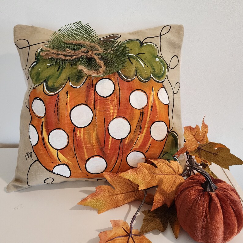 Puede incluir: Una almohada beige con un dise&ntilde;o de calabaza pintado a mano. La calabaza es naranja con lunares blancos y hojas verdes. La almohada est&aacute; adornada con un lazo de arpillera marr&oacute;n.