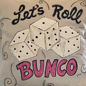 Peut inclure: Un coussin beige avec un motif peint &agrave; la main repr&eacute;sentant trois d&eacute;s blancs &agrave; points noirs et les mots "Let's Roll Bunco" en noir et rose. Des volutes d&eacute;coratives et des paillettes entourent les d&eacute;s et le texte.